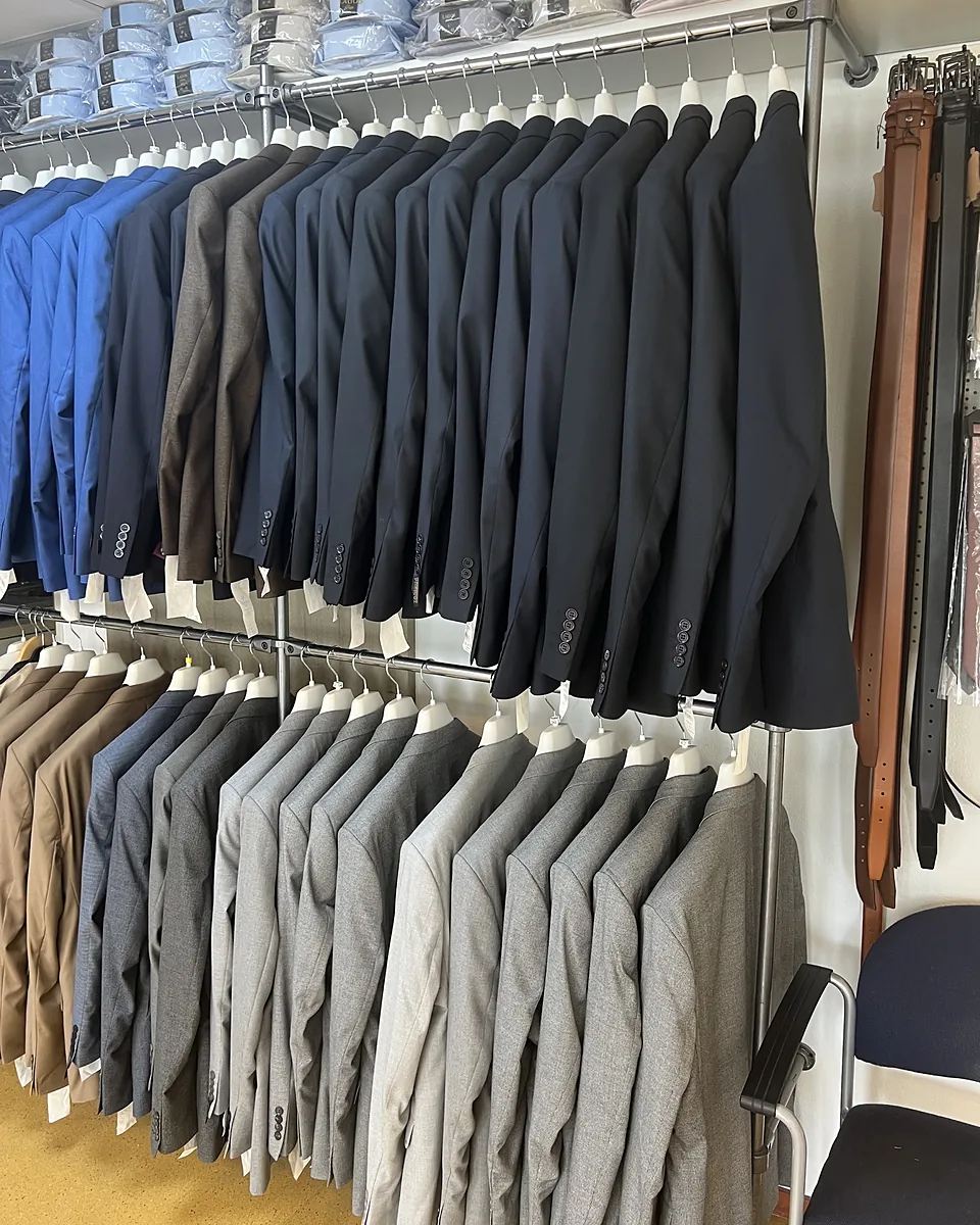 Een rek met colberts in blauw, grijs en zand, overhemden erboven en riemen opzij